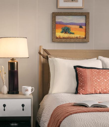 A cozy bedroom with a neatly made bed, a decorative pillow, an open book, a nightstand with a lamp, a mug, dried flowers, and a framed landscape painting on the wall above the headboard.
