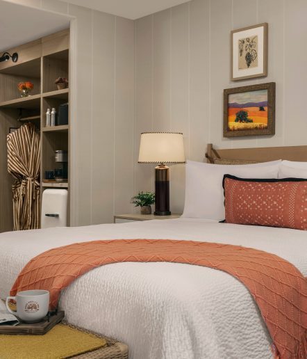 A cozy, modern bedroom with a neatly made bed topped with a rust-colored pillow and throw, a bedside table with a lamp and plant, open closet with a striped robe, wall art, and a mug on a woven bench in the foreground.