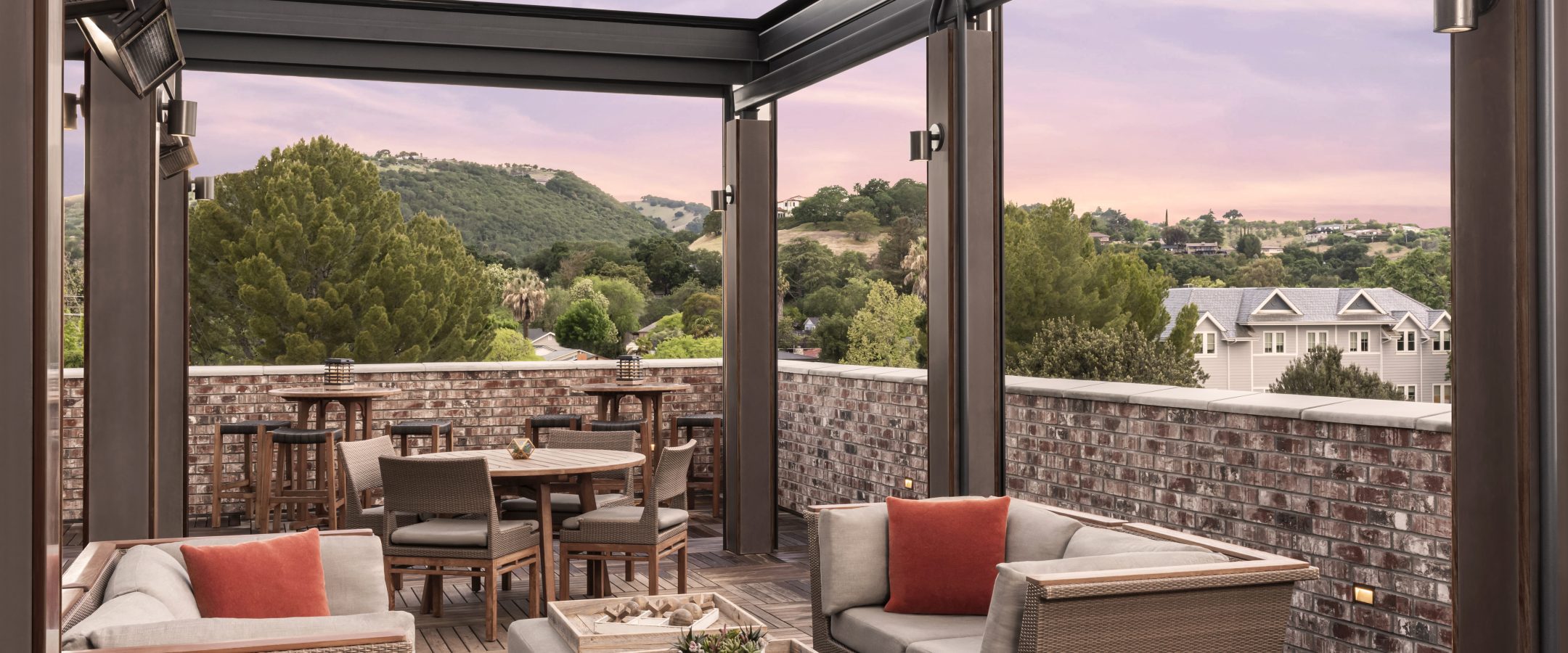 Outdoor patio with modern wicker furniture, cushioned seats, and a table under a wooden pergola. Several tables and chairs are in the background, with hills and houses visible beyond a brick wall at sunset.