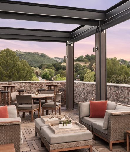 An outdoor patio with cushioned sofas, wooden tables, and chairs overlooks green hills under a pink and purple sky. Brick walls and a pergola frame the relaxed, modern seating area.