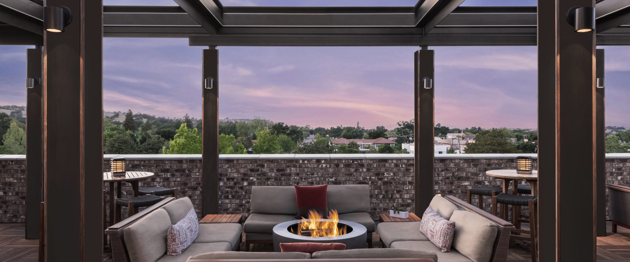 Modern rooftop lounge with cushioned seating around a fire pit under a pergola at sunset, overlooking trees and rooftops with a colorful sky in the background.
