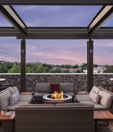 Rooftop patio with cushioned seating around a round fire pit, wooden tables, and a partial glass roof. Trees and buildings are visible beyond the brick wall at sunset with a purple sky.