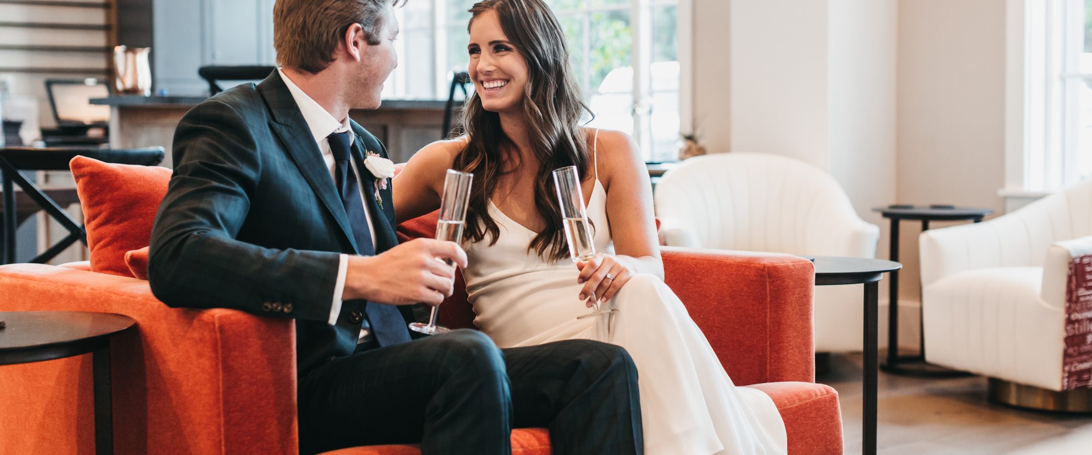 A man in a suit and a woman in a white dress sit close together on an orange couch, smiling at each other and holding champagne glasses in a bright, modern lounge.