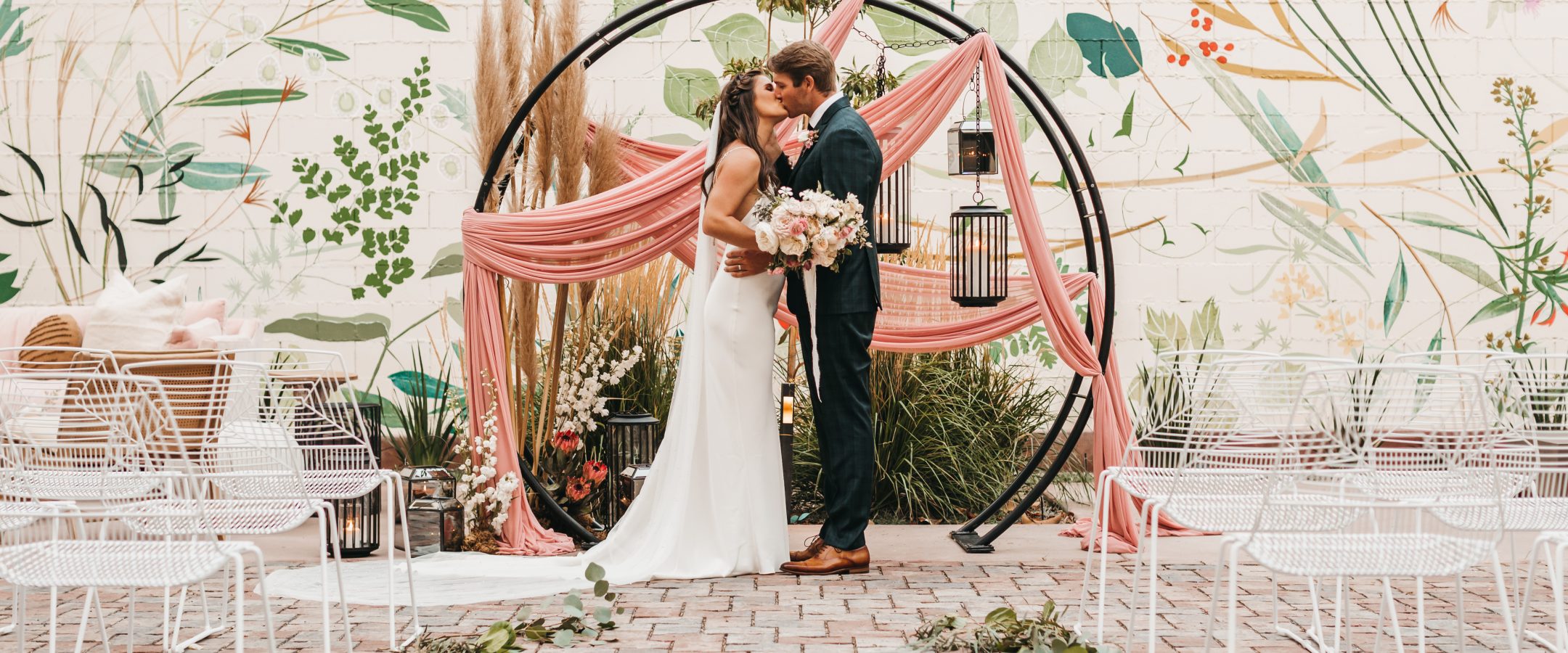 A bride and groom kiss in front of a circular wedding arch decorated with pink fabric and greenery, surrounded by white chairs and a wall mural featuring large botanical designs.