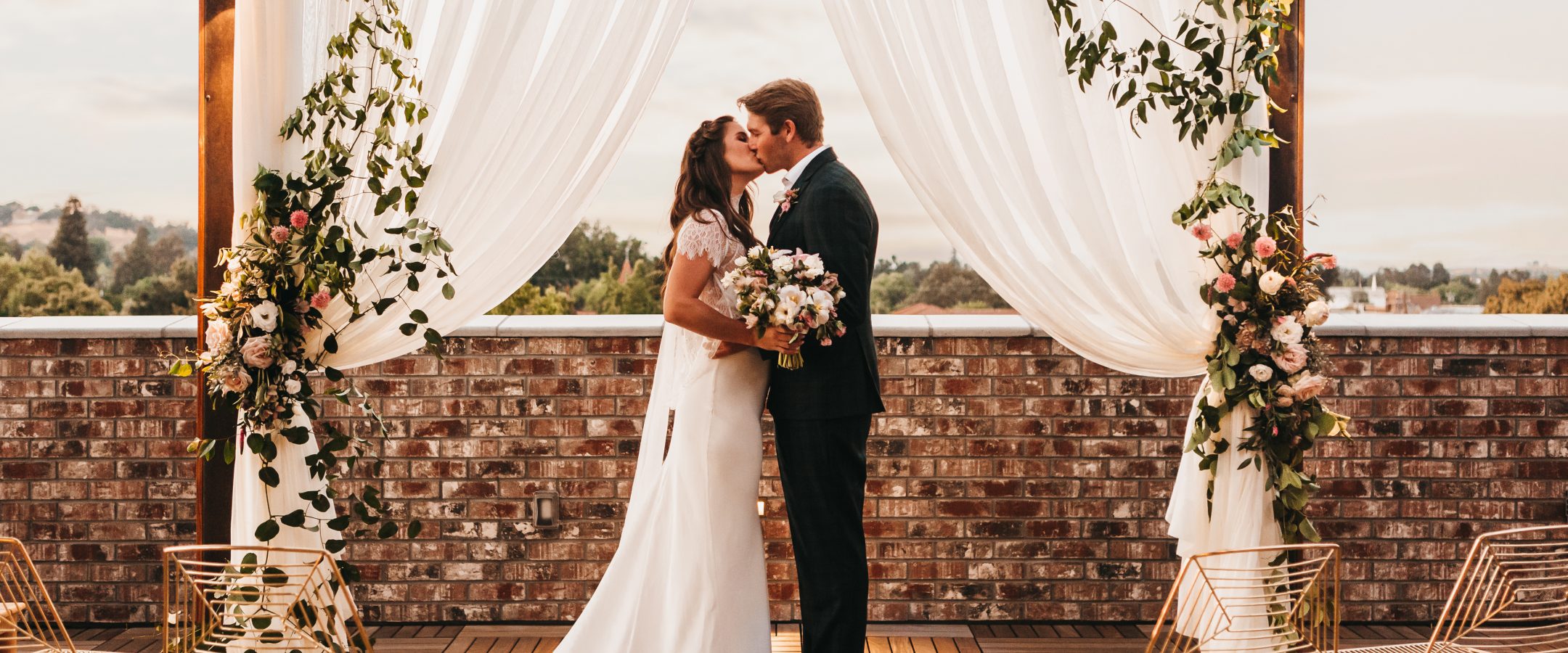 A bride and groom kiss under a white draped arch decorated with greenery and flowers on an outdoor wooden deck, with empty gold chairs arranged on both sides and a brick wall in the background.
