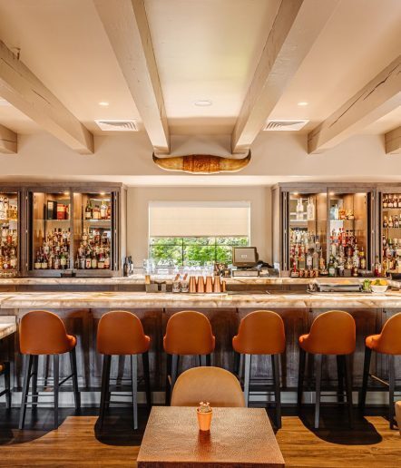 A stylish bar with tan leather stools, a polished wooden counter, shelves stocked with bottles, copper shakers, and a decorative longhorn skull mounted above the bar. A window lets in natural light behind the bar.