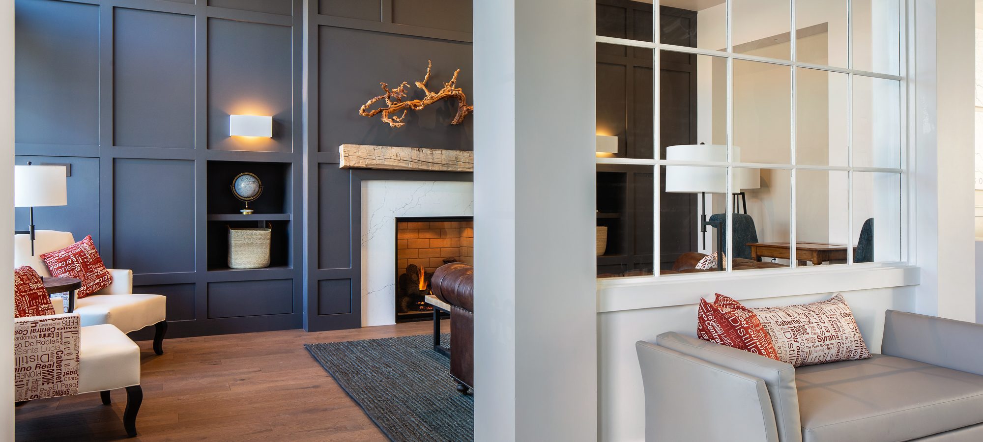 Modern living room with dark paneled accent wall, fireplace, white armchairs with red pillows, gray loveseat, hardwood floors, and large window with grid design separating spaces. Warm, cozy and elegantly decorated.
