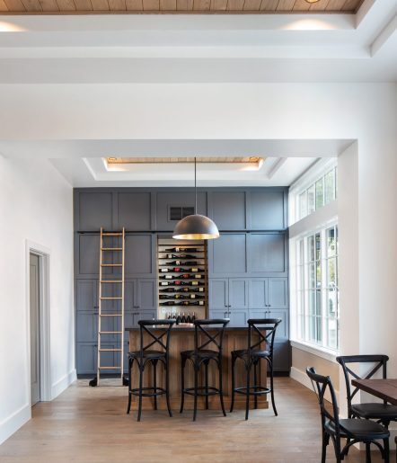 A modern home bar features navy paneled walls, a wine rack, a wooden ladder, a wooden bar with four black chairs, a large dome pendant light, and large windows letting in natural light.