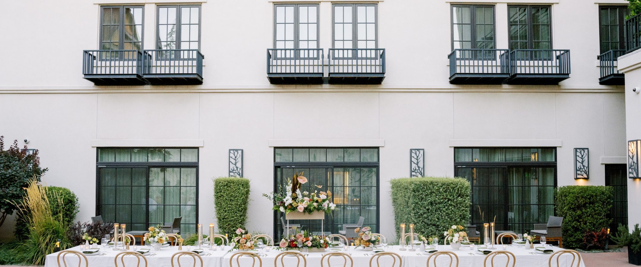 A long, elegant outdoor dining table with light wooden chairs, white tablecloth, and floral centerpieces is set in a courtyard in front of a modern building with large windows and small black balconies.