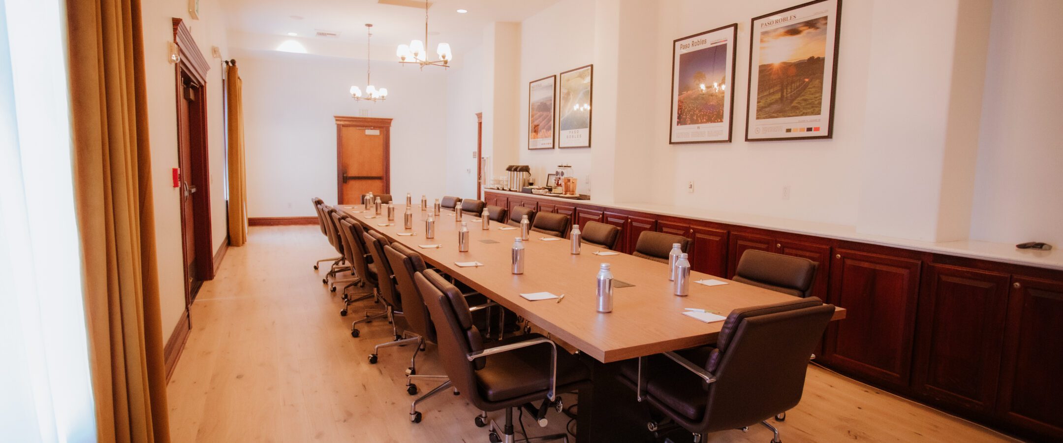A modern conference room with a long wooden table surrounded by black chairs, water bottles placed at each seat, and framed posters on the white walls. The room is well-lit with chandeliers and natural light.