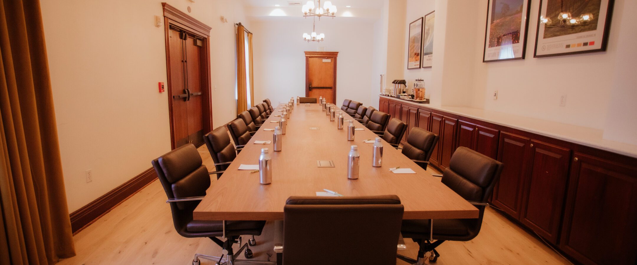 A modern conference room with a long wooden table, black office chairs, bottled water, notepads, and framed posters on the wall. The room has wood cabinets, chandeliers, and natural light from windows.