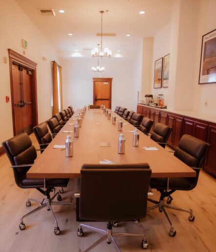 A long, wooden conference table with black chairs arranged around it in a spacious, well-lit meeting room. Water bottles and notepads are set at each seat; refreshments and artwork are visible along one wall.