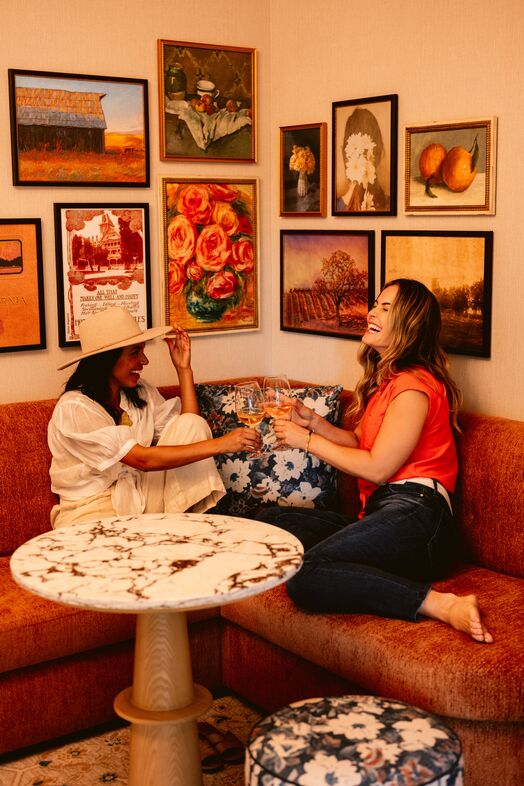 Two women sit on a rust-colored couch, smiling and clinking wine glasses. One wears white and a hat; the other wears a red top and jeans. Behind them is a wall filled with colorful framed art and paintings.