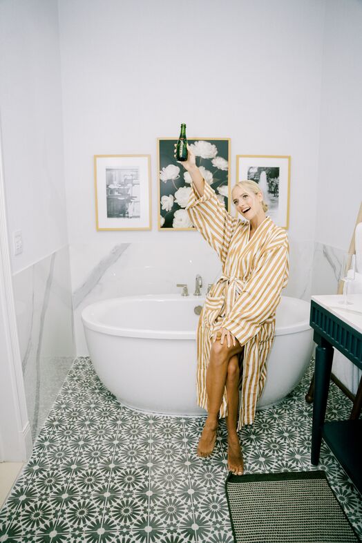 A smiling person in a yellow and white striped robe sits on the edge of a white bathtub, holding up a bottle. The bathroom has patterned floor tiles, framed wall art, and a dark blue vanity.