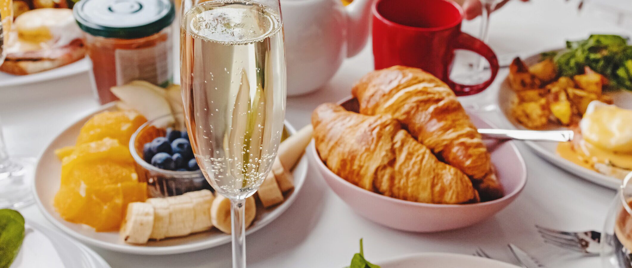 A festive breakfast table features a glass of sparkling wine, croissants, fresh fruit, eggs, salad greens, red mugs, and a white teapot—bringing the inviting charm of a Steakhouse Paso Robles brunch to your morning.