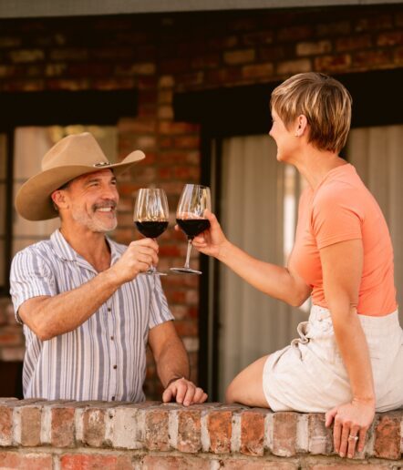 A smiling man in a cowboy hat and a woman in an orange shirt clink glasses of red wine while sitting on a brick wall outside a building.