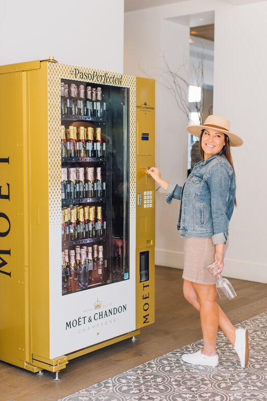 A smiling woman in a hat and denim jacket stands beside a gold Moët & Chandon champagne vending machine at The Piccolo at Paso Robles Inn, holding a champagne flute and preparing to make a selection.