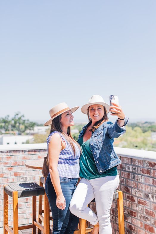 Two women wearing hats stand on The Piccolo at Paso Robles Inn’s rooftop patio. One takes a selfie while the other smiles, surrounded by bar stools and a brick wall under a clear blue sky.