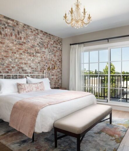 A bright bedroom in a boutique hotel in Paso Robles features a brick accent wall, a white bed with blush accents, a cushioned bench, chandelier, and large sliding glass doors opening to a balcony with views of the trees.