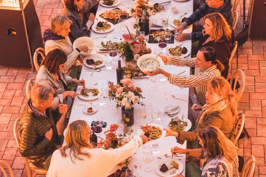 A group of people sit around a long, elegantly set outdoor table at The Piccolo at Paso Robles Inn, sharing food and enjoying a meal together, surrounded by flowers and wine on a brick patio.