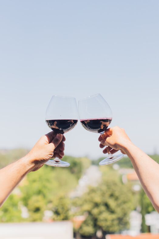 Two hands holding glasses of red wine, clinking them together for a toast outdoors at The Piccolo at Paso Robles Inn, with a blurred background of trees and blue sky.
