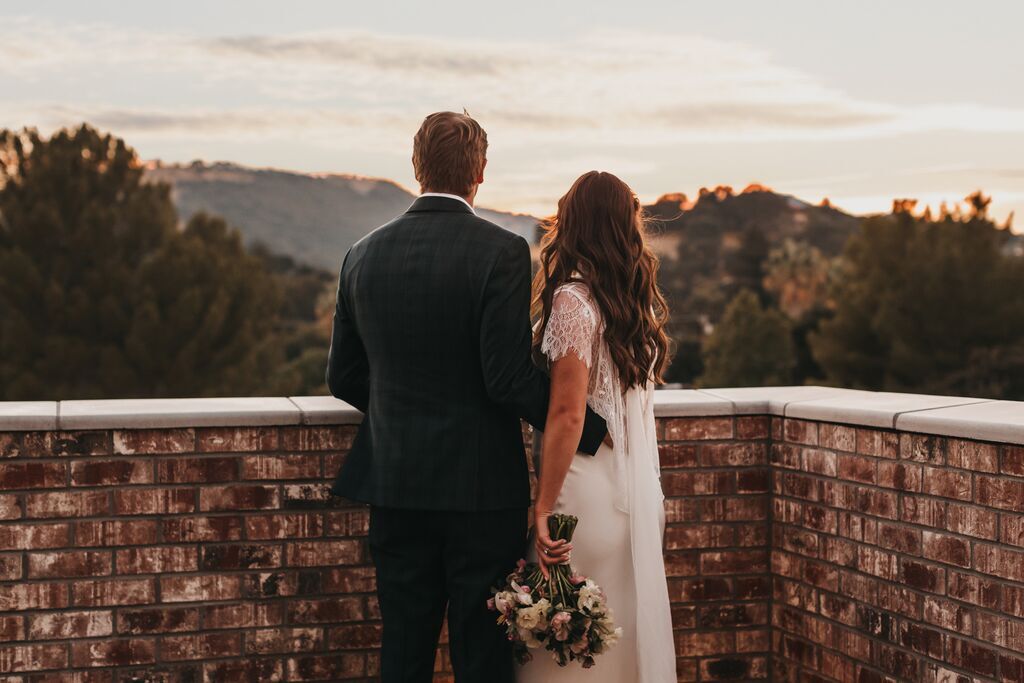 A bride and groom stand side by side on a brick terrace at The Piccolo at Paso Robles Inn, facing a scenic sunset over distant hills. The bride holds a bouquet of flowers behind her back.