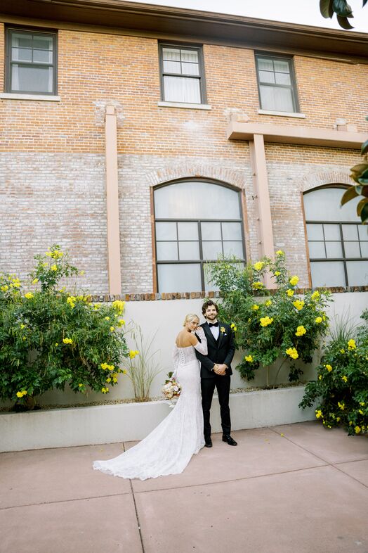 A bride in a white lace gown and a groom in a black tuxedo stand together at The Piccolo at Paso Robles Inn, framed by large windows, yellow flowering bushes, and classic brickwork. The bride holds a bouquet and leans her head on the groom’s shoulder.