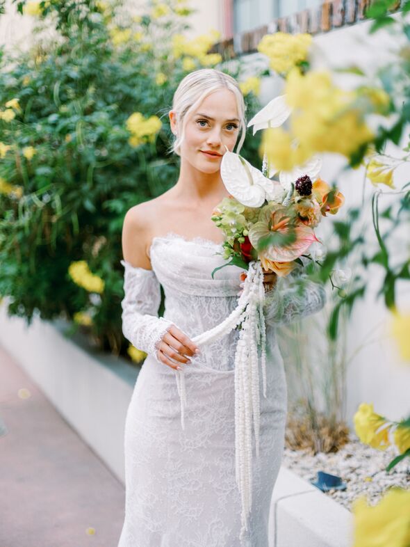 A bride in an off-the-shoulder white lace wedding dress holds a colorful bouquet while standing outdoors amid green foliage and yellow flowers at The Piccolo at Paso Robles Inn.