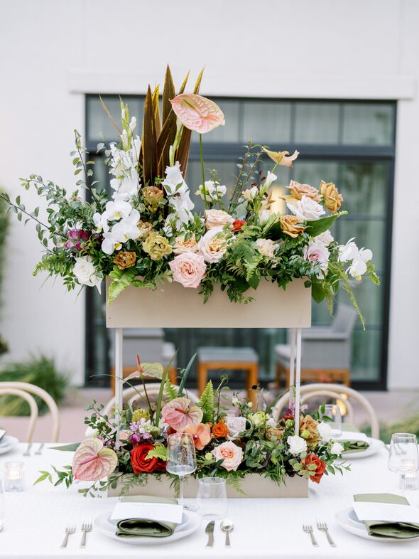 A table centerpiece at The Piccolo at Paso Robles Inn features two tiers of vibrant floral arrangements with roses, orchids, and greenery on a white stand, surrounded by neatly arranged table settings.