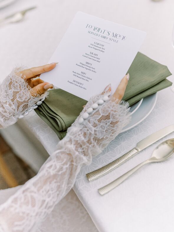 A person wearing lace sleeves holds a printed dinner menu above a green napkin and white plate on an elegantly set table with silver cutlery at The Piccolo at Paso Robles Inn.