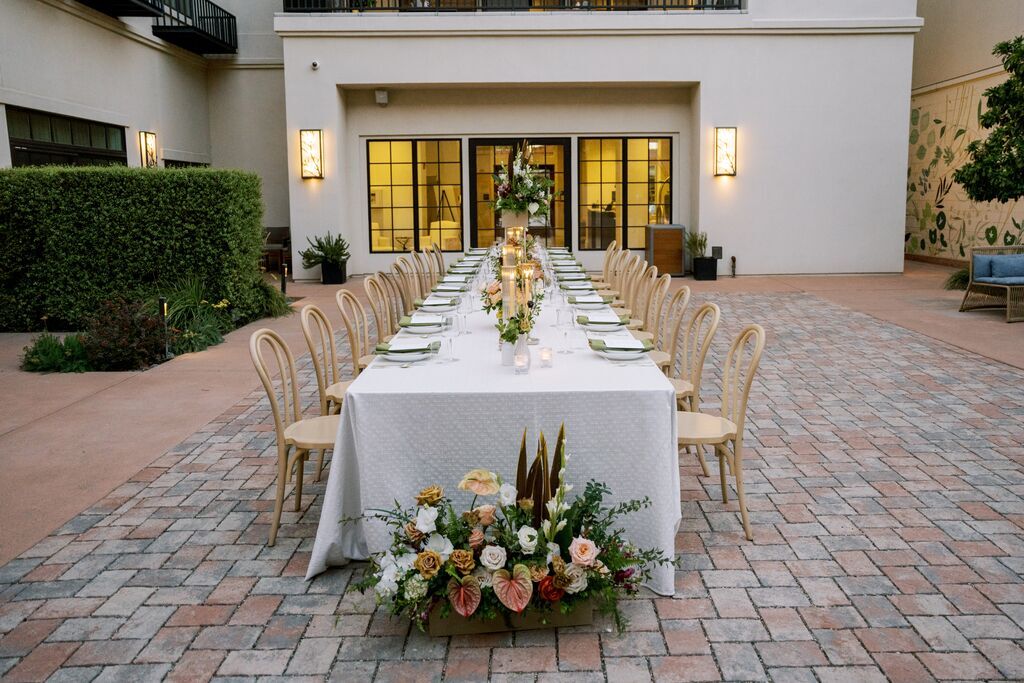 A long, rectangular outdoor dining table with a white tablecloth is set for a formal event at The Piccolo at Paso Robles Inn. Wooden chairs line both sides, floral centerpieces adorn the table, and a modern building frames the courtyard setting.