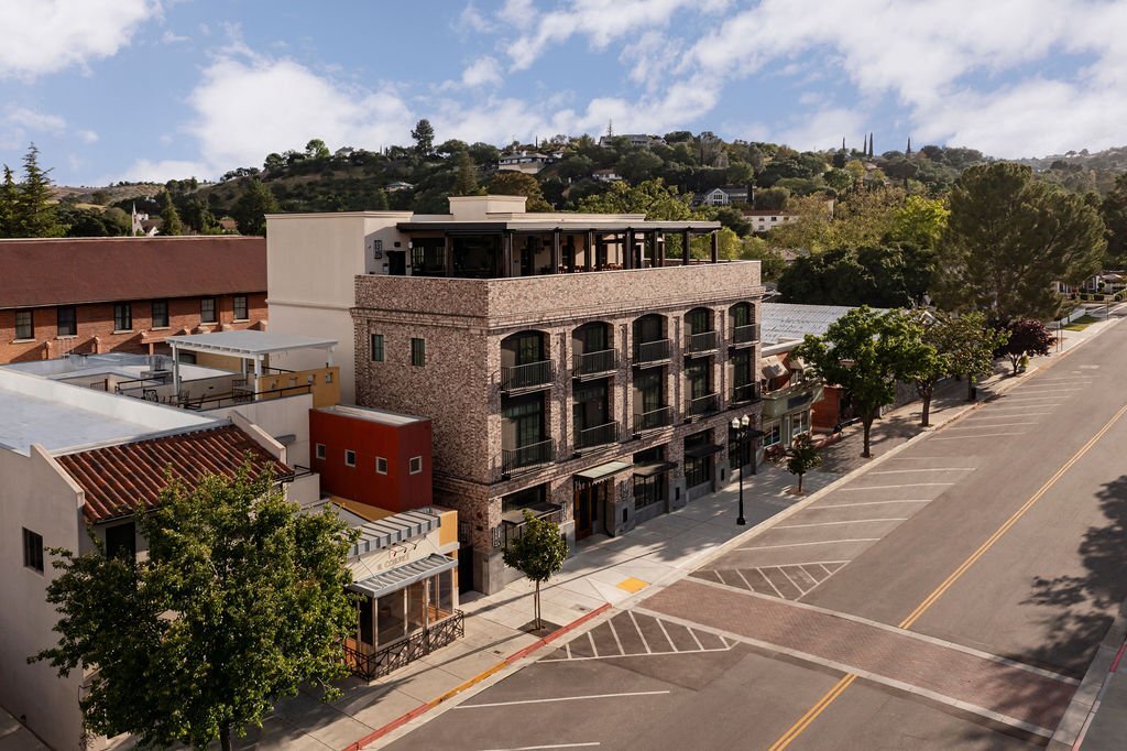 A modern brick building with balconies and rooftop seating stands on a quiet street lined with trees, with hills and greenery in the background under a partly cloudy sky.