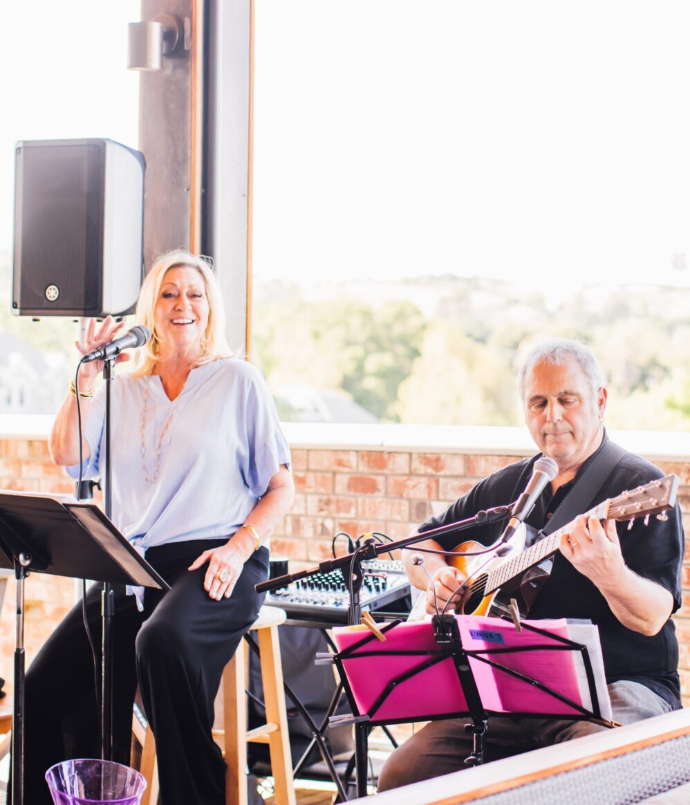 A woman sings into a microphone and a man plays an acoustic guitar beside her. They are performing together outdoors, with music stands and sound equipment visible around them. Both appear to be enjoying the performance.