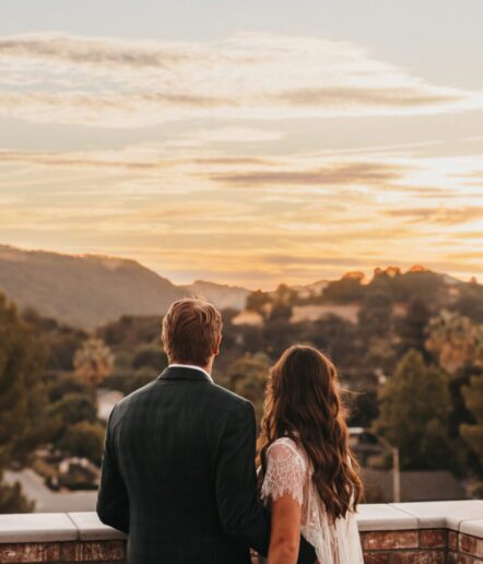A couple stands close together, facing away and looking at a scenic sunset over rolling hills and trees, with soft clouds in the sky.