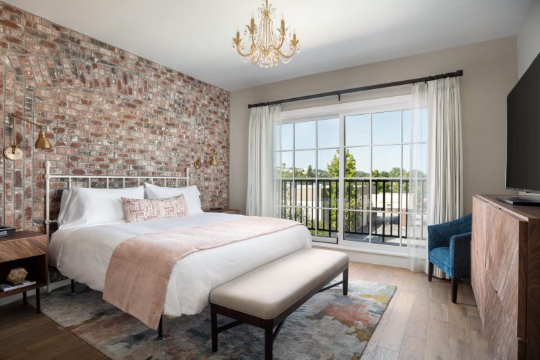 A stylish bedroom features a brick accent wall, a large bed with white and pink bedding, a bench at the foot, a chandelier, wooden furniture, a blue chair, and a window with sheer curtains overlooking greenery.