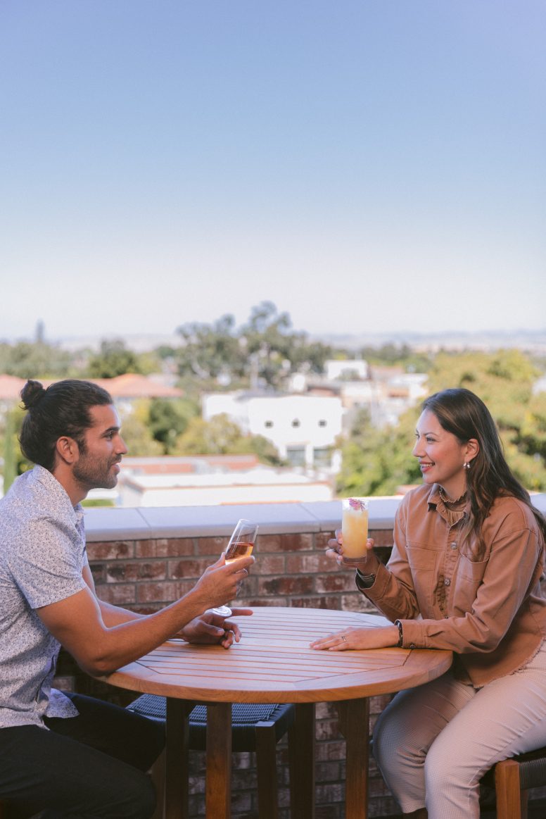 A man and woman sit across from each other at an outdoor table, smiling and holding drinks, with a cityscape and trees visible in the background under a clear sky.