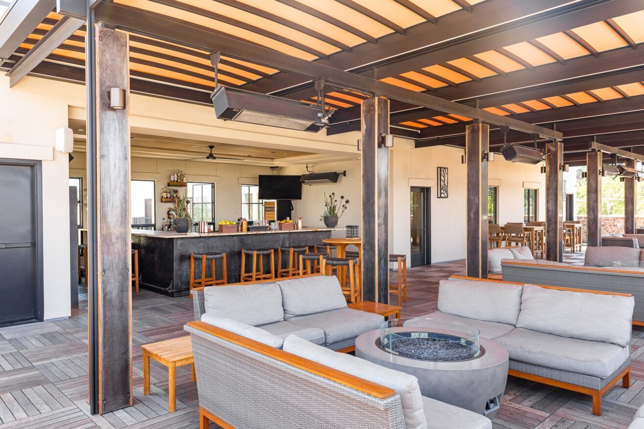 Modern outdoor patio with cushioned seating around a fire pit, wooden tables and chairs, and a bar with stools in the background under a pergola-style roof. The setting appears bright and inviting.