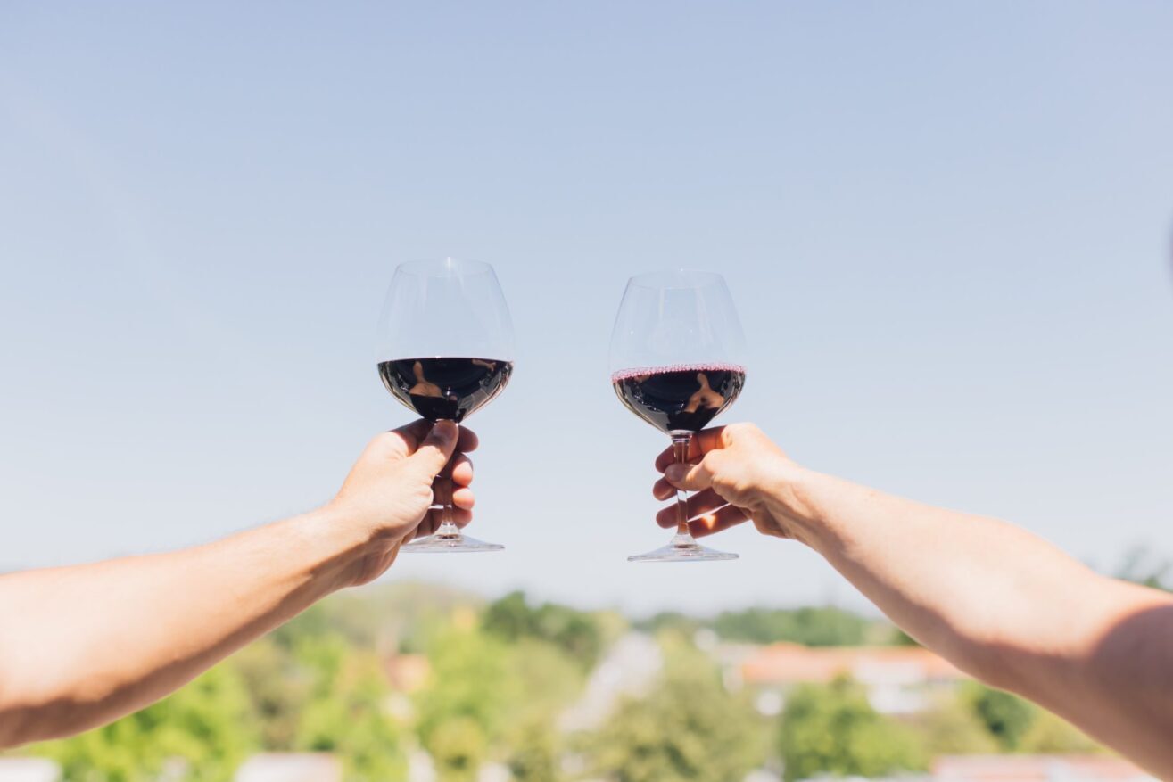Two hands holding red wine glasses are raised in a toast against a clear blue sky, with blurred green trees and rooftops in the background.