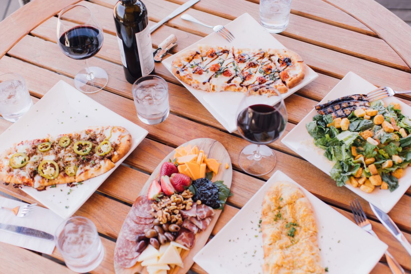 A wooden table set with flatbreads, a cheese and charcuterie platter, Caesar salad, glasses of red wine, water glasses, and a bottle of wine.