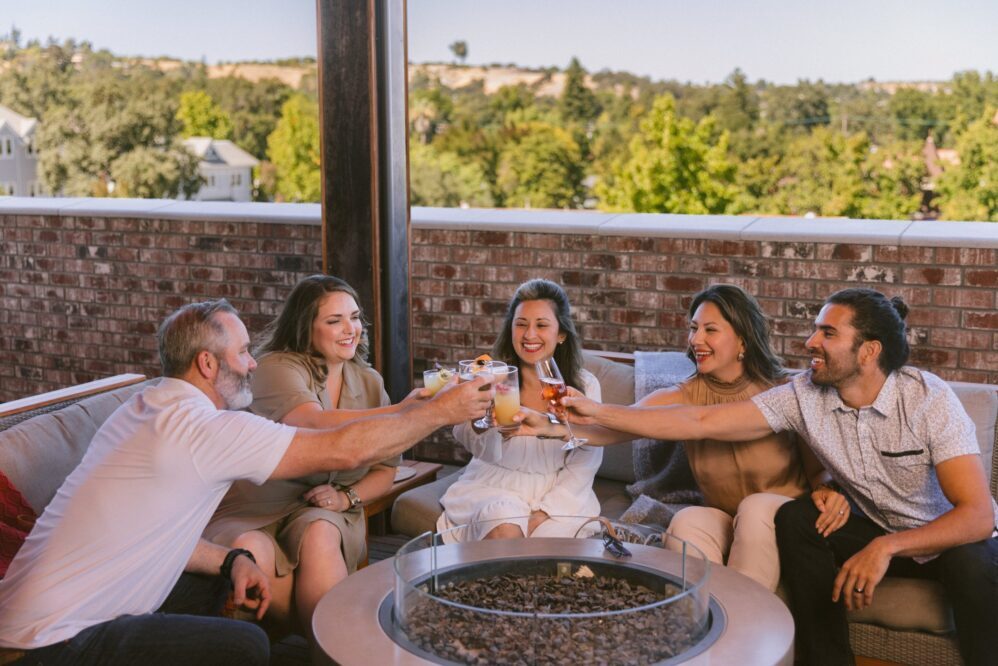 Five people sitting on an outdoor patio sofa, smiling and clinking drinks together around a fire pit table, with trees and houses in the background on a sunny day.