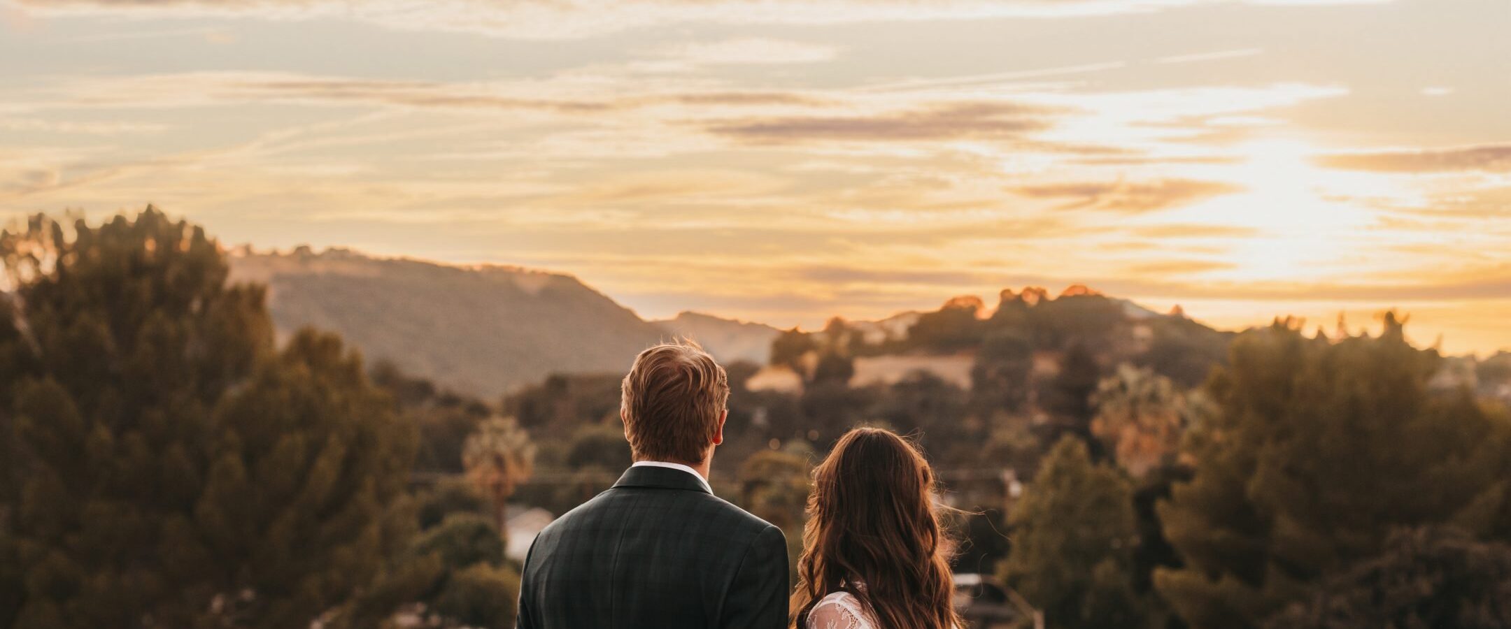 A couple stands side by side on a balcony, looking at a scenic sunset over rolling hills and trees. The sky is partly cloudy with warm, golden light illuminating the landscape.