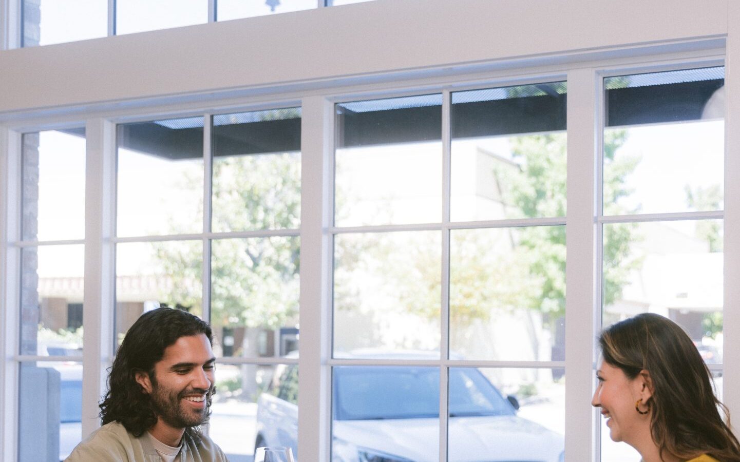 Two people sit at a wooden table by large windows, smiling and talking while tasting wine. Several wine glasses and a plate of snacks are on the table, with daylight streaming into the modern room.