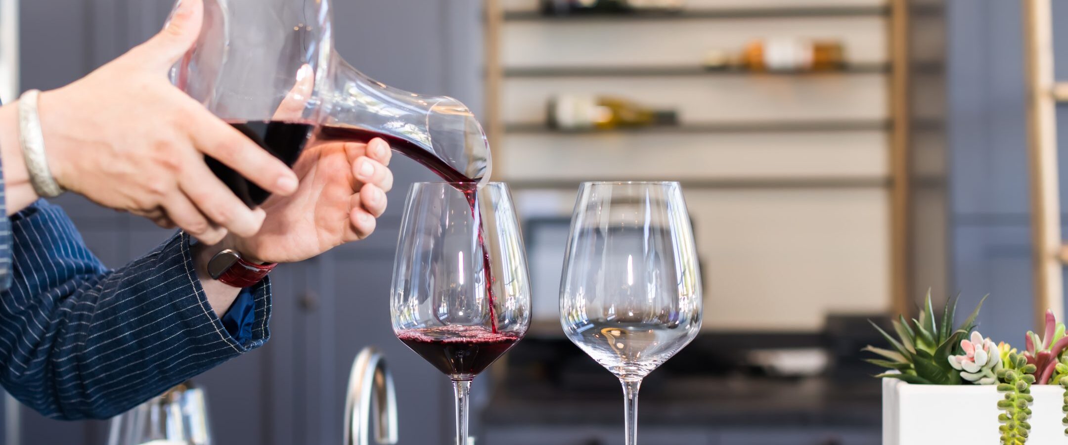 A person pours red wine from a decanter into a wine glass on a counter, with another empty glass beside it. Shelves with wine bottles and a small potted plant are in the background.