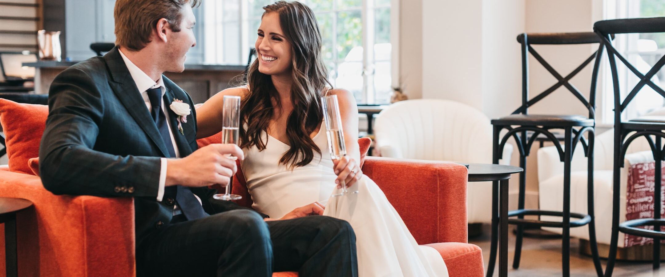 A smiling couple in formal attire sit close together on a red couch, holding champagne glasses and looking at each other warmly in a bright, modern room with large windows.