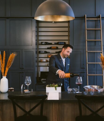 A man in a suit pours red wine into a glass at a modern wine bar with dark cabinets, a large dome pendant light, a ladder, and decorative vases with dried grasses. Bottles of wine are displayed on shelves behind him.