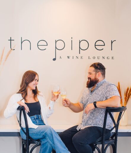 A woman and a man sit on chairs clinking wine glasses and smiling at each other in front of a wall that reads the piper A WINE LOUNGE. Dried plants decorate the sides of the scene.