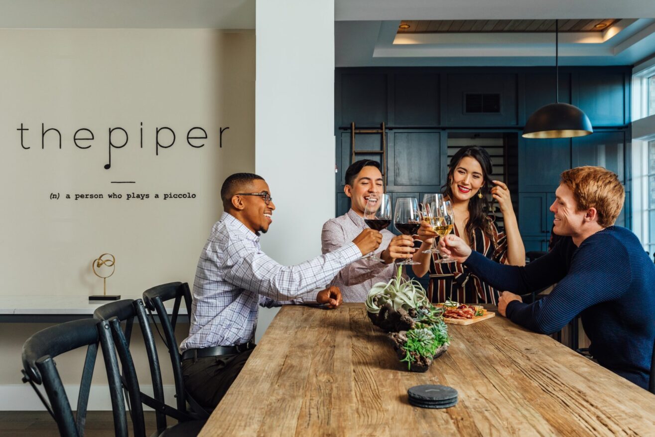 Four people sit around a wooden table in a modern kitchen, smiling and clinking glasses together. There is food on the table and a sign on the wall reads the piper.