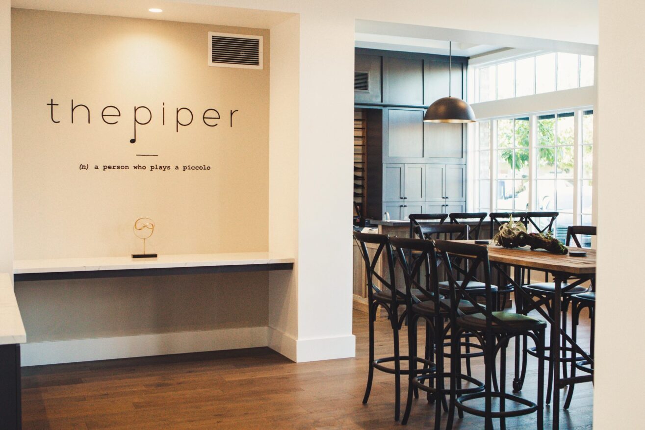 Modern dining area with wooden tables and chairs, large windows, and a wall with the text the piper (n): a person who plays a piccolo. Natural light fills the stylish, minimalist space.