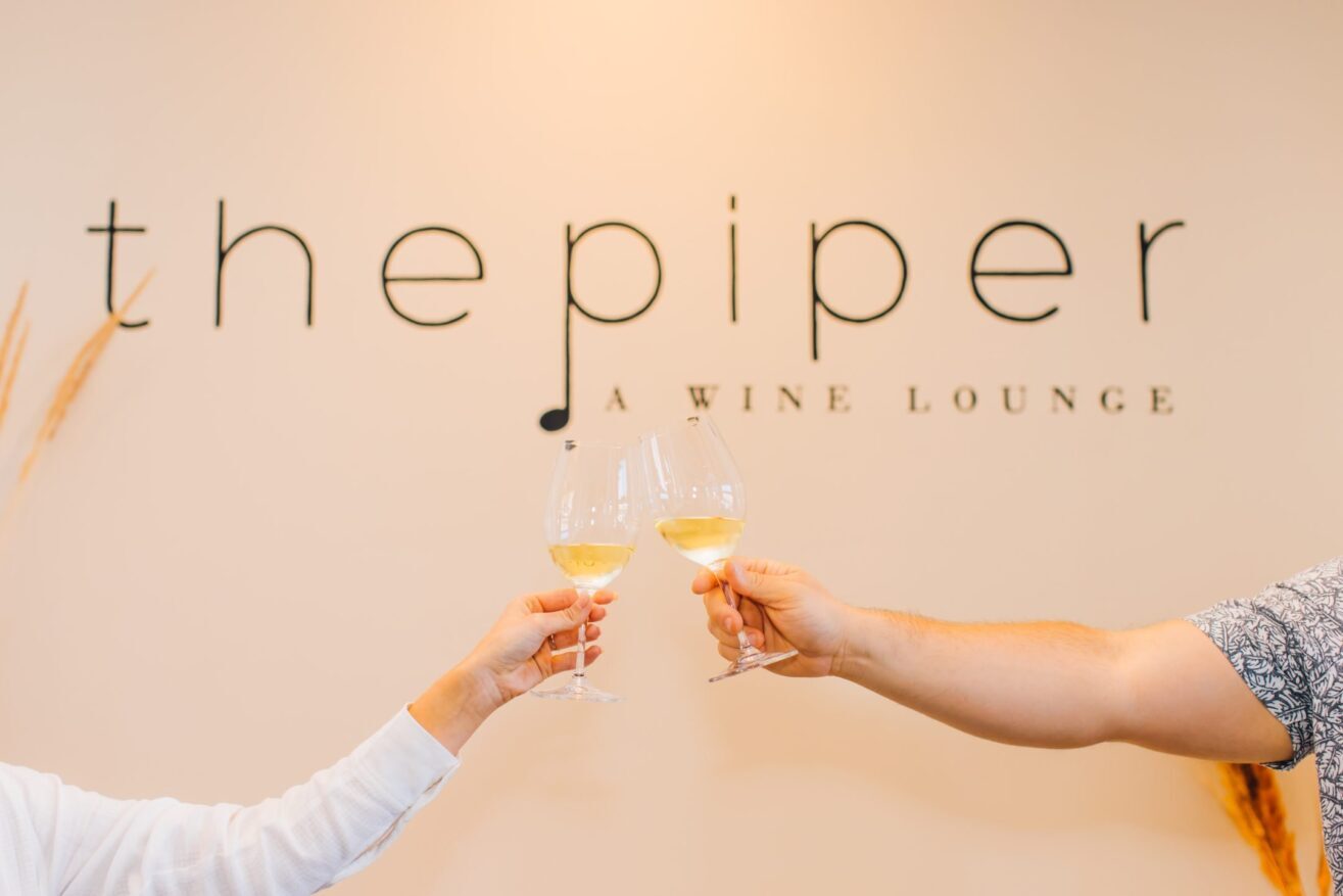 Two people clink glasses of white wine in front of a wall sign that reads the piper A WINE LOUNGE. Only their hands and arms are visible.