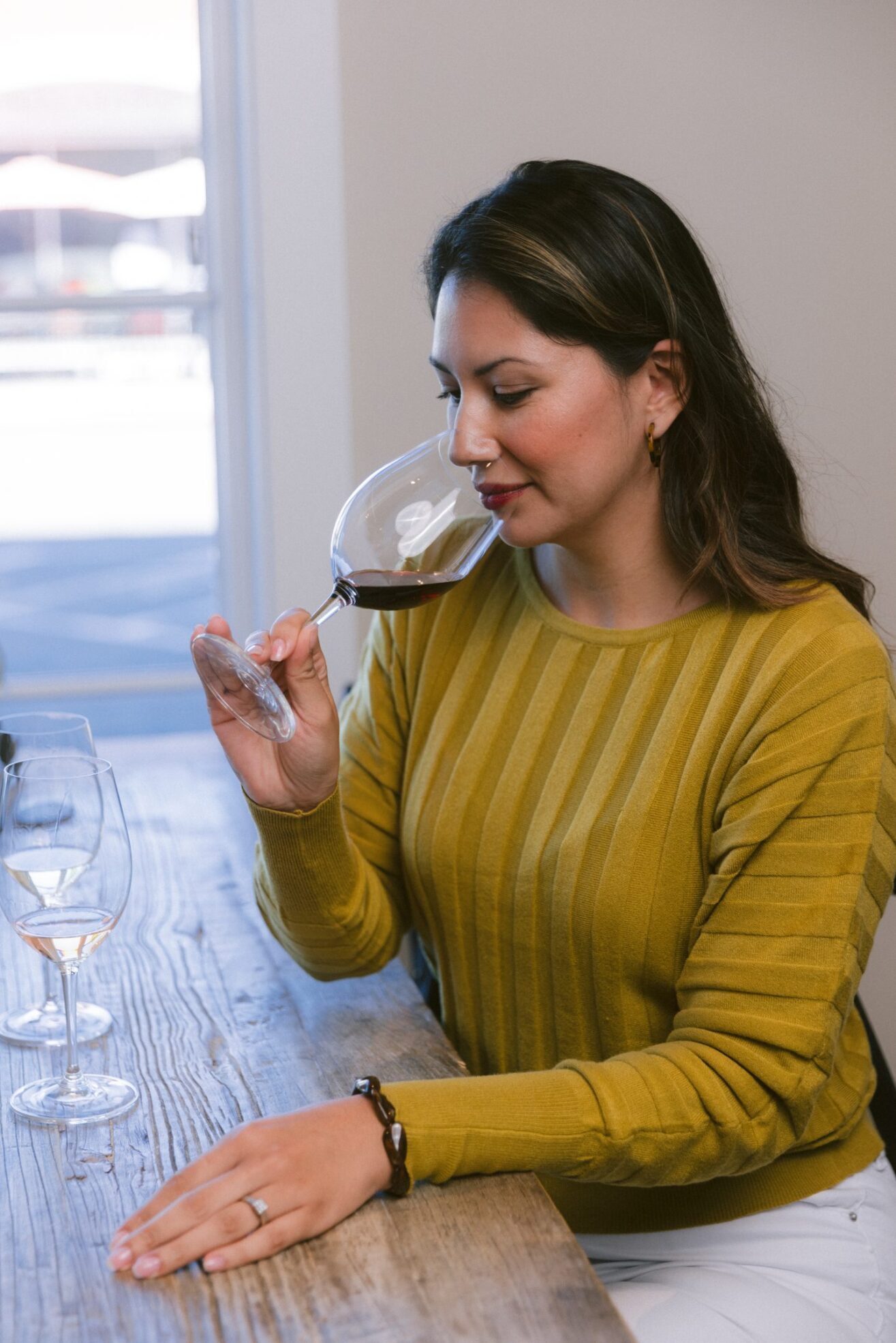 A woman wearing a yellow sweater sits at a table, holding a glass of red wine up to her nose and smelling it. Another glass with white wine is on the table beside her.
